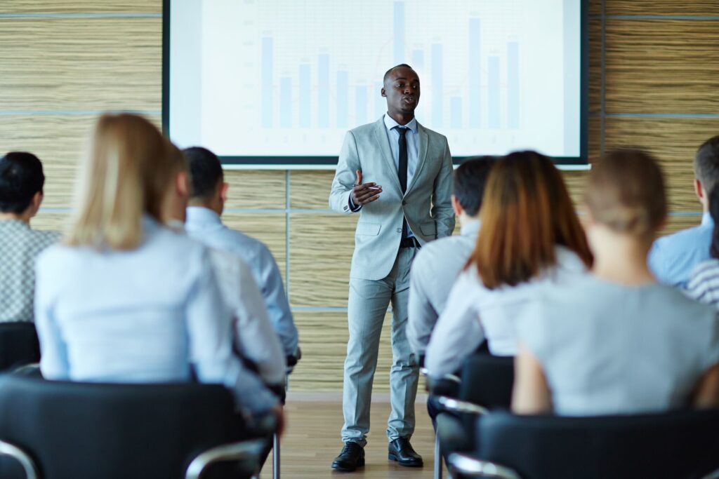 A professional speaker presents to an audience, highlighted by a data chart on a screen. This setting emphasizes business communication and analysis.