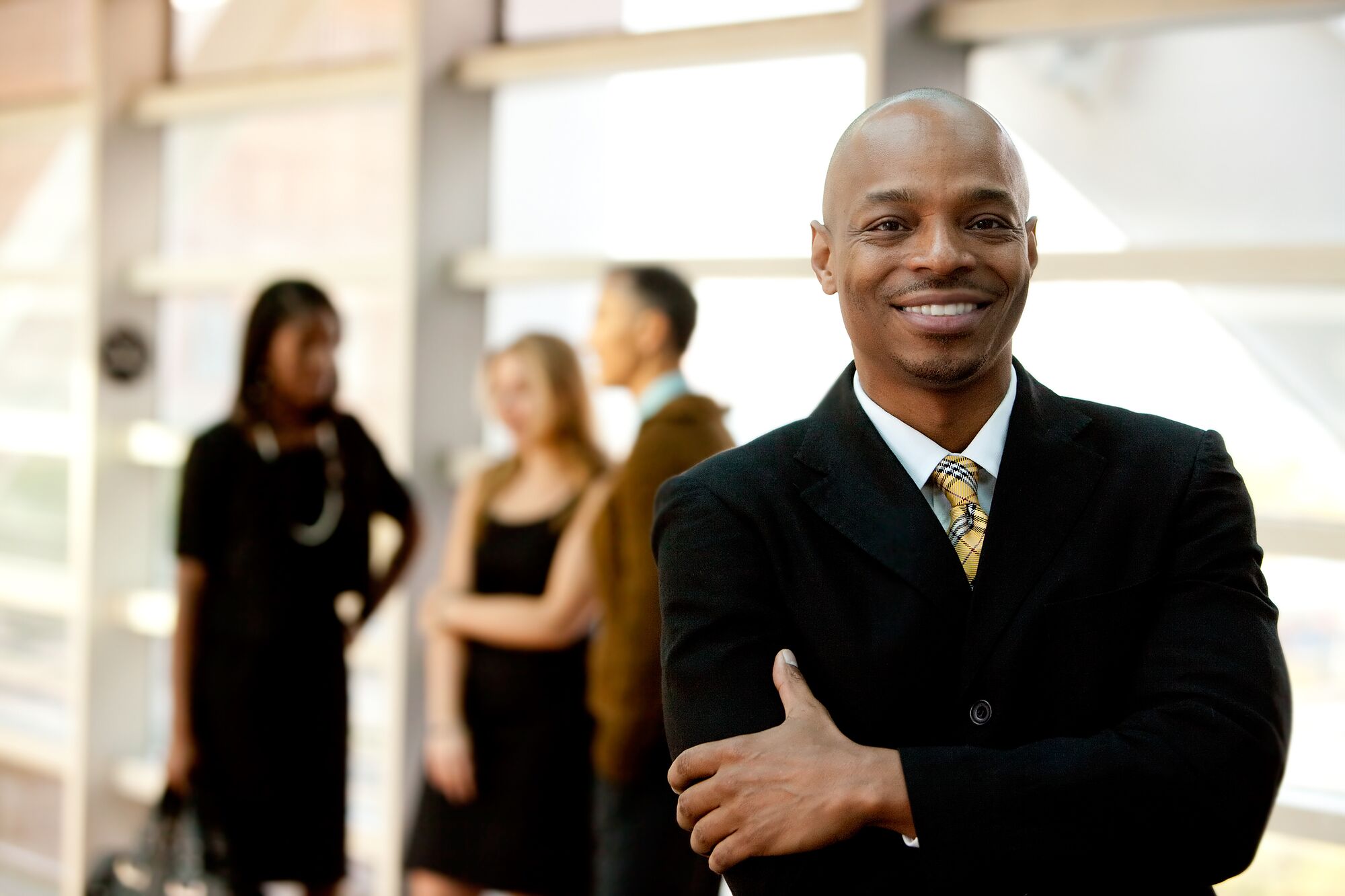 A confident man in a black suit smiles while four people converse in the background, highlighting a professional networking event.