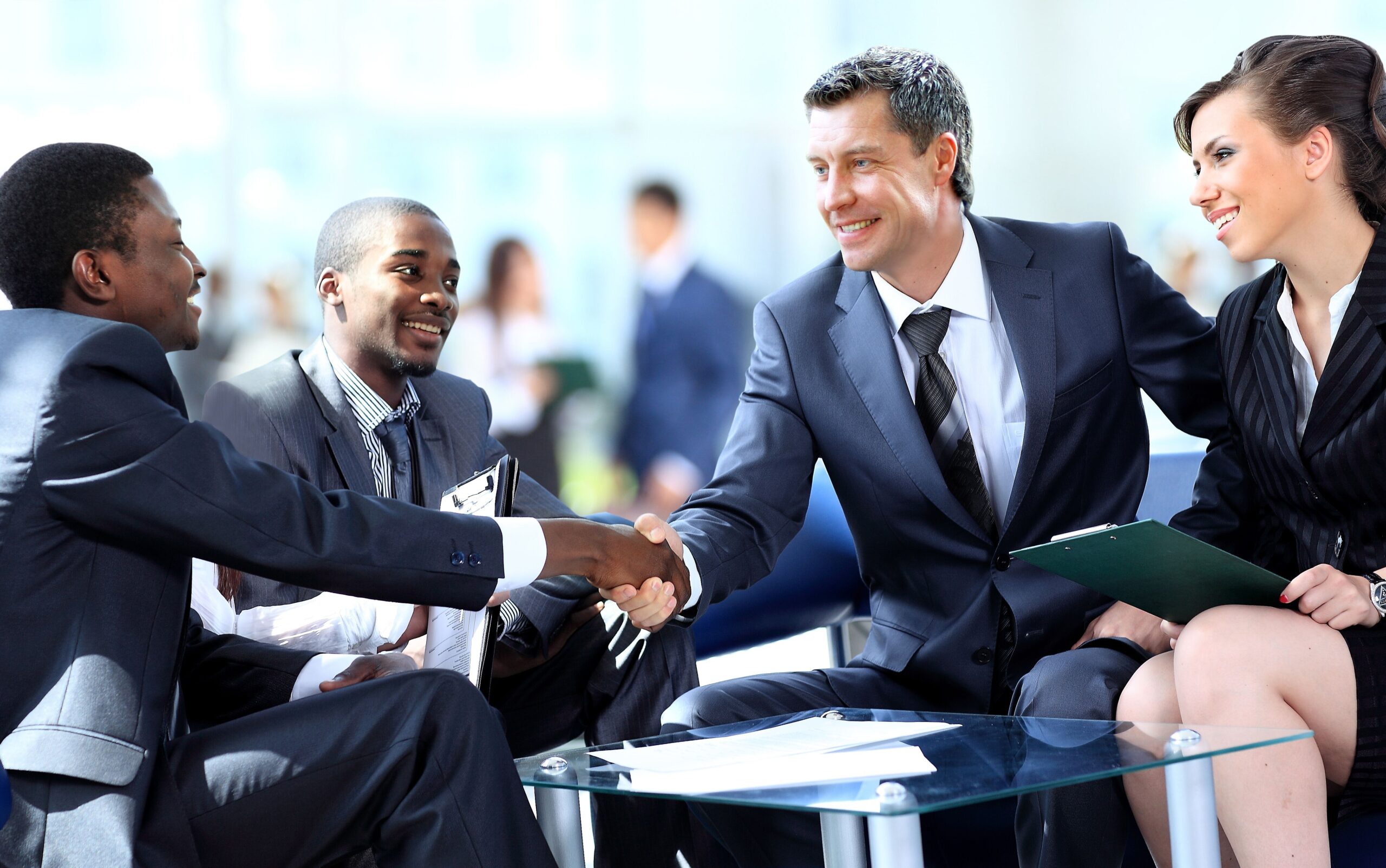 Four business professionals in suits sit around a glass table, smiling and engaged.