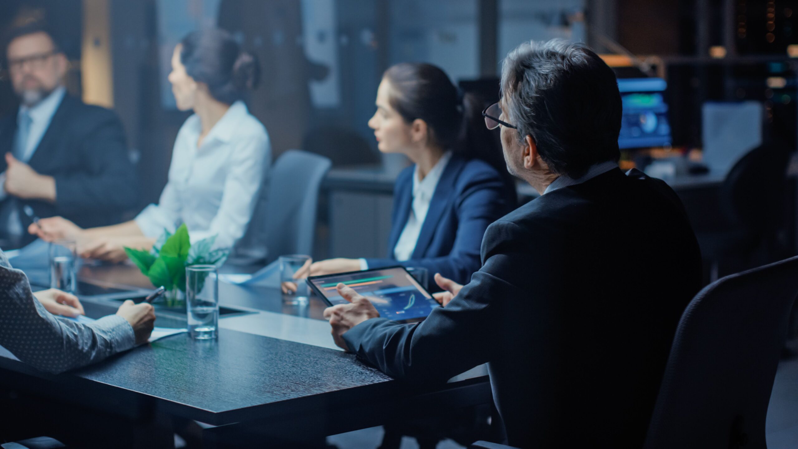 A group of business professionals in formal attire are engaged in a serious discussion around a conference table, with laptops and documents present.