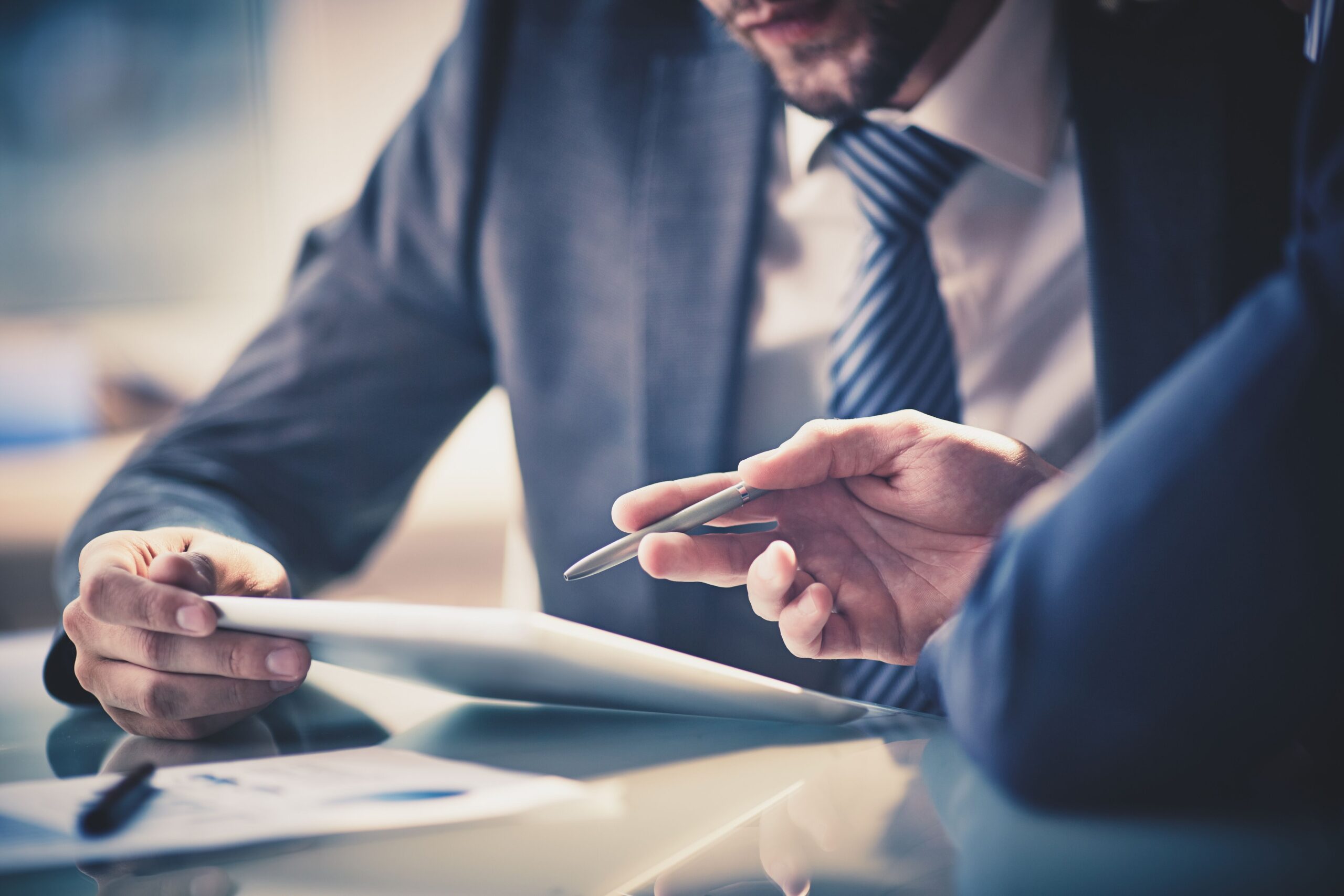 Two business professionals in suits discuss a document on a tablet at a glass table, creating a focused and collaborative atmosphere.