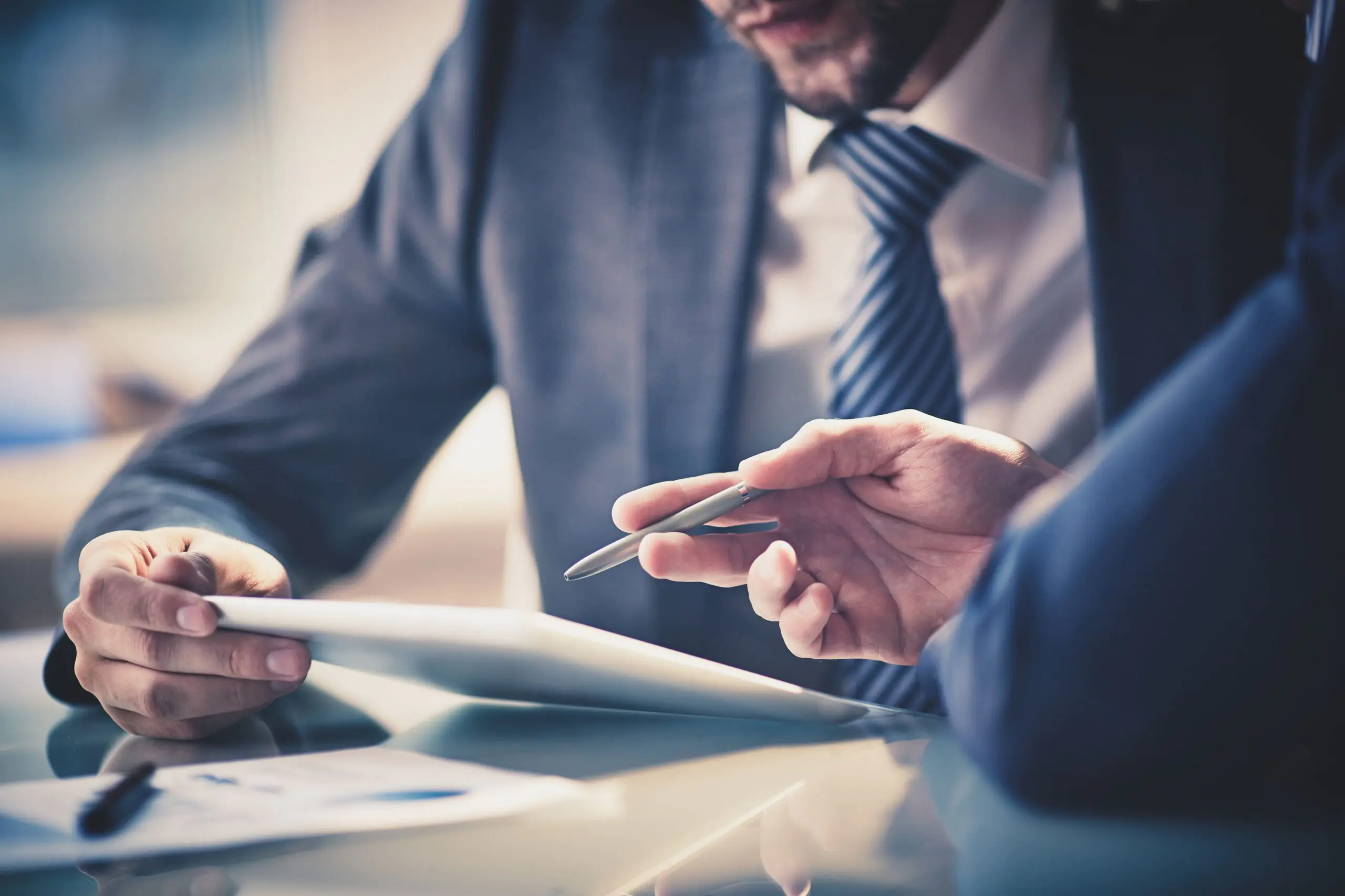 Two business professionals in suits discuss a document on a tablet at a glass table, creating a focused and collaborative atmosphere.