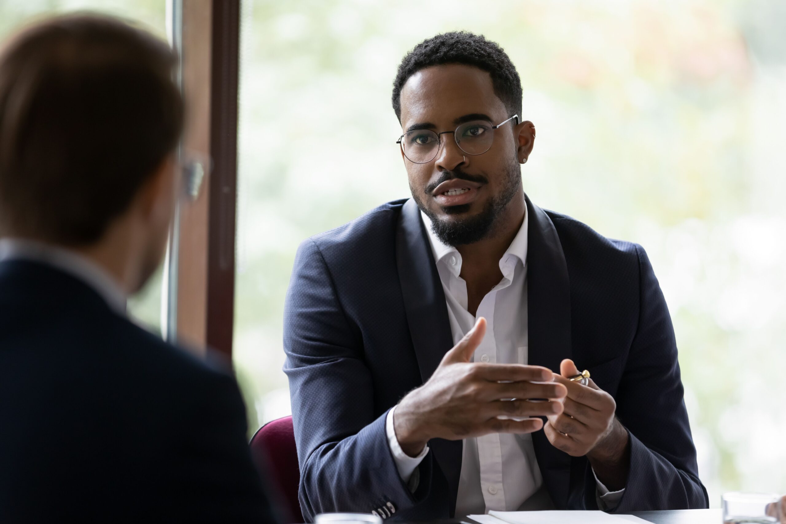 A man in glasses and a suit speaks thoughtfully in an office setting, gesturing with his hands.