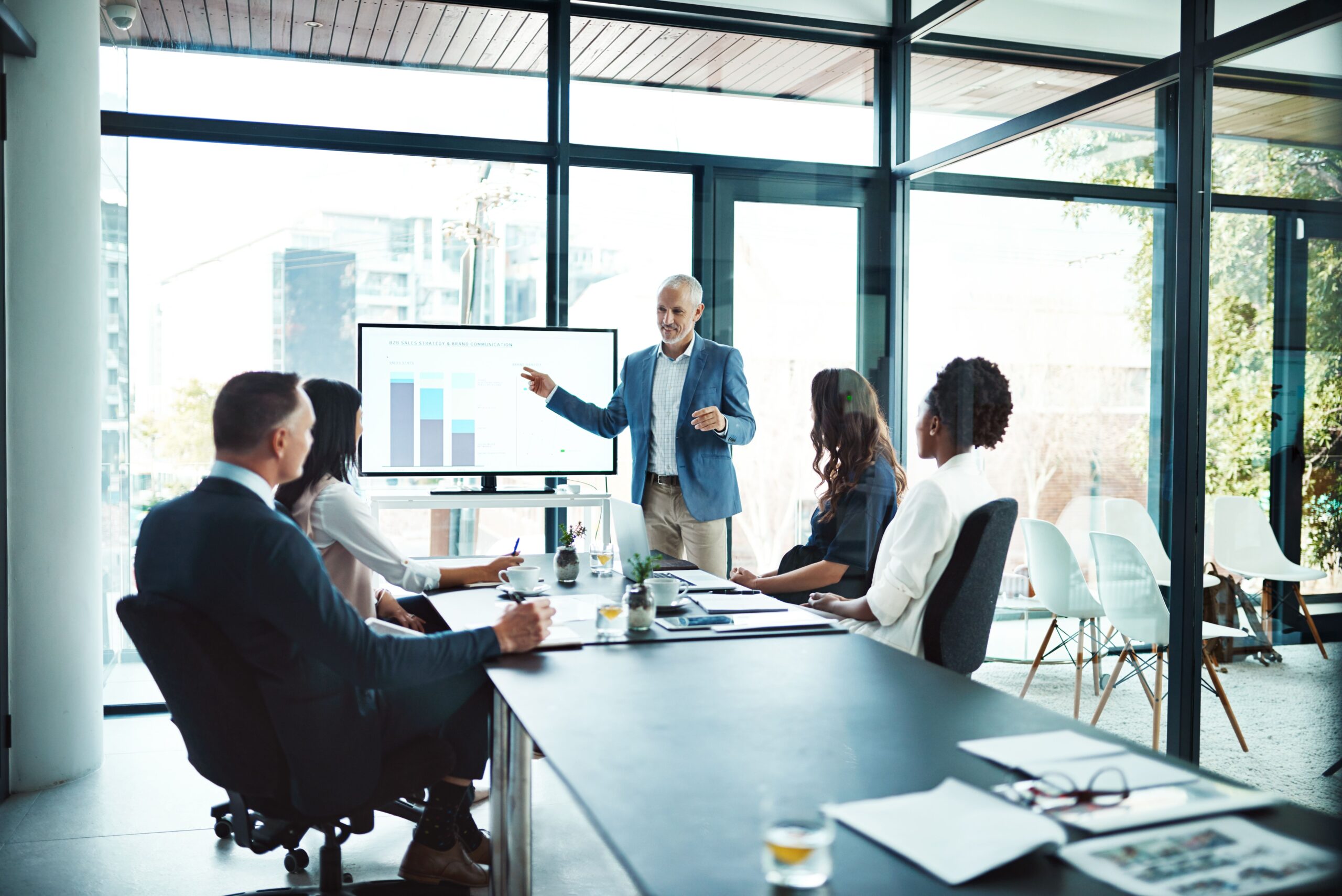 A man in a blazer presents a bar chart on a screen to a diverse group seated in a bright conference room, creating a professional and focused atmosphere.