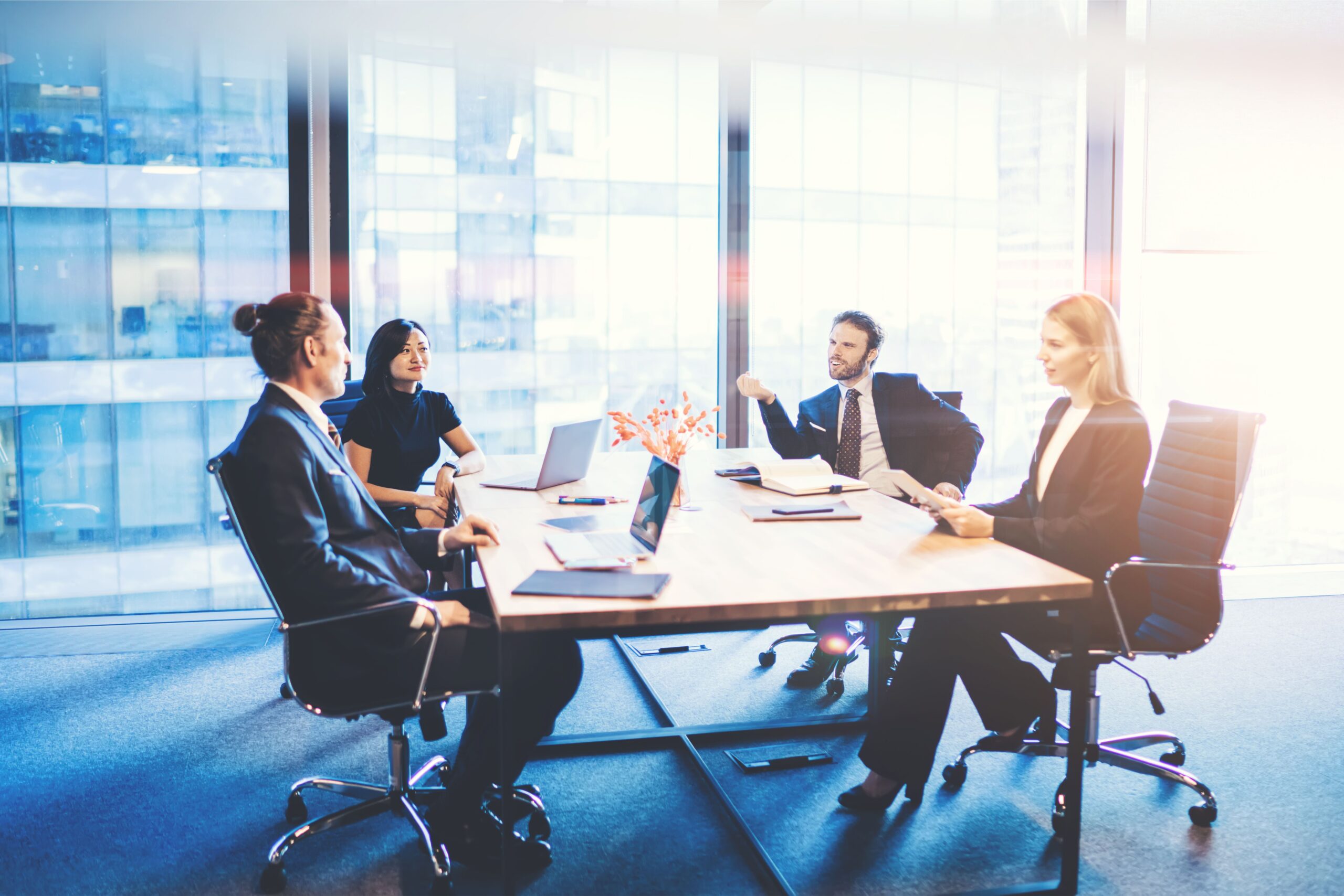 A diverse group of four professionals in business attire sit around a conference table in a glass-walled office, engaged in a meeting.